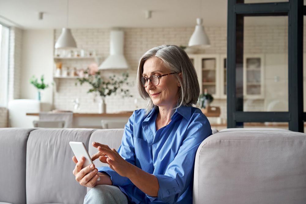 Eine Person sitzt auf einem hellen Sofa und hält ein Smartphone in der Hand. Im Hintergrund ist eine moderne Küche mit Pflanzen und hellen Möbeln zu sehen.